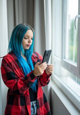 Vertical portrait of a beautiful blue-haired hipster Caucasian woman standing in front of the window taking a selfie with her mobile phone. Using technology. clipart