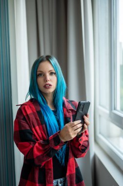 Pencerenin önünde durup cep telefonuyla selfie çeken güzel mavi saçlı beyaz bir kadının dikey portresi. Kameraya bakıyorum. Teknoloji kullanılıyor.