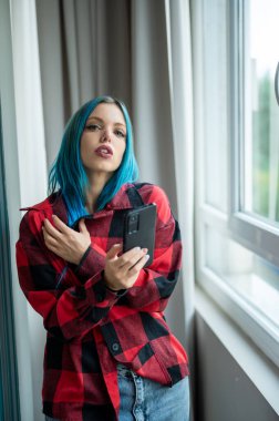 Vertical portrait of beautiful blue haired hipster caucasian woman standing in front of window taking selfie with her mobile phone. Looking at camera. Using technology. clipart