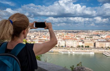 Budapeşte, Macaristan, 29 Ağustos 2022. Orta yaşlı bir kadın cep telefonuyla tepenin üstündeki muhteşem şehir manzarasının fotoğraflarını çekiyor. Mavi gökyüzü ve beyaz bulutlarla güzel bir yaz günü.