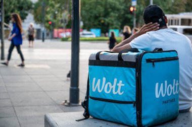 Budapeşte, Macaristan, 30 Ağustos 2022. Wolt 'ta bir işçi, yemek dağıtım şirketi. Açık mavi teslimat çantası ve logosu yanında oturarak mola veriyor. Gig ekonomisi.