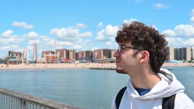 New York City, ABD, 21 Ağustos 2024. Genç beyaz bir adam Coney Island rıhtımına bakıyor ve cep telefonuyla selfie çekiyor. Teknoloji kullanarak yaşam tarzı.