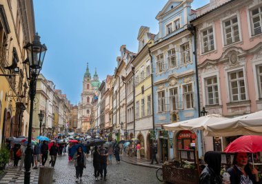 Prag, Çek Cumhuriyeti, 5 Ağustos 2023. Mala Strana Caddesi 'ndeki sokak fotoğrafçılığı, arka planda St. Nicholas Kilisesi' nin çan kulesi. Yağmurlu bir gün, şemsiyeli insanlar. Seyahat yerleri.