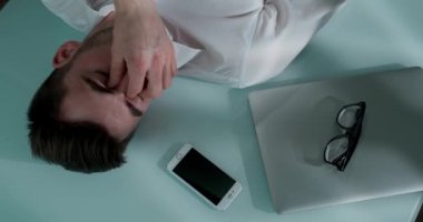 Sad man lying on the table with laptop, smartphone and glasses. He closes his eyes and covers his face with his hands. Man is alarmed and thoughtful. Camera view from above.