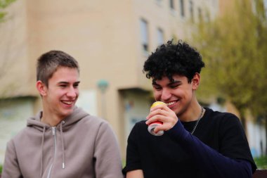 İki genç adam bankta içki içiyor, şehir hayatı tarzında, dışarıda arkadaşları var.