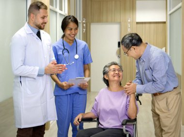 Beyaz önlüklü beyaz doktor ve steteskoplu genç hemşire test sonuçlarını hasta ve ailesine bildirin. Tekerlekli sandalyedeki yaşlı bir kadın kocasıyla mutlu bir şekilde gülümsüyor..