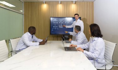 Hastanedeki çeşitli bölümlerin müdürü yıllık toplantıya katıldı. Toplantı lideri sunumlar için düz ekran TV kullanır. Herkesin kendi dizüstü bilgisayarı var..