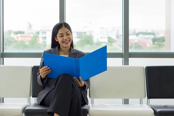 Young asian businesswoman in gray suit sit on a bench near the window, reading document in blue folder.