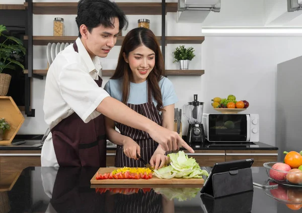 Young asian man dressed in an apron pointing at cooking recipe on tablet computer. His girlfriend  holding knife slices the vegetable into pieces on a wooden chopping board. Atmosphere in a kitchen.