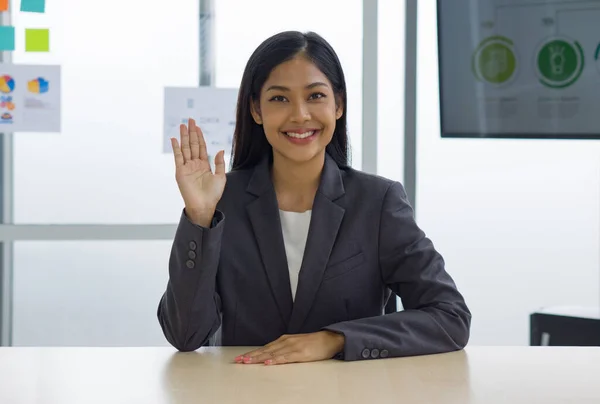 stock image Young asian female employee in gray suit sit relax with smile face, waving hand in front while sit at her desk. There was a note paper and document on glass board in the background.