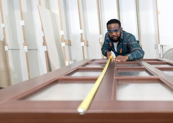 Short black hair man with moustache and beard use a tape measure to measure the size of a wooden door. Plywood storage compartment are on the background. Carpenter worker work in a furniture factory.