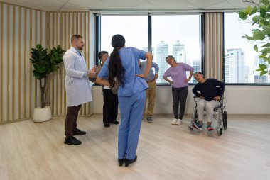 Bir grup yaşlı insan, Kafkasyalı doktor ve genç Asyalı fizyoterapist ile sabah egzersizi yapıyor. Huzurevinde sabah atmosferi.