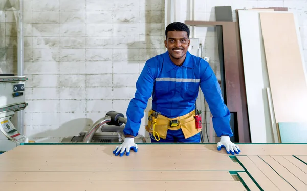 Young technician in blue mechanic jumpsuit, protective glove and yellow Tool Belts loading plywood on working table. Production line atmosphere in a wooden furniture factory.