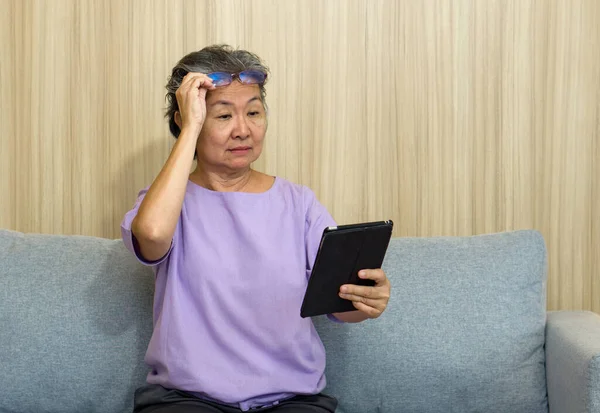 Asian retired elderly with eyeglasses, sitting on the sofa watching at tablet computer. With presbyopia syndrome she cannot focus on nearby objects.