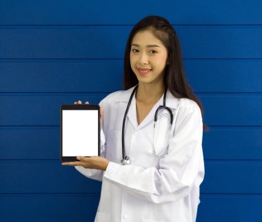 Young asian doctor in white gown and stethoscope holding white screen tablet computer, standing in front of blue wall.