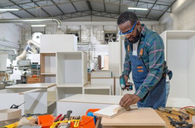 Short black hair man with moustache and beard use electric drill to drill the planks as they were prepared at the wooden furniture factory. A desk full of hand tools.