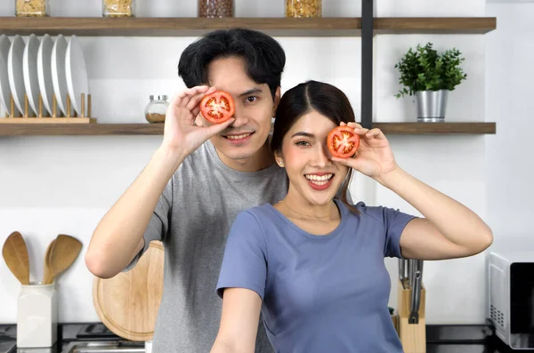 Young couple in casual clothe, hold slice tomato in eye position. Asian lover spend morning time together in the kitchen.