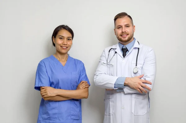 Young caucasian doctor and Asian  physical therapist stand with arm cross in front of white wall.