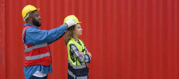 A foreman with mustache and beard helps a female apprentice wear a hardhat. Both of them wearing reflective safety vest. A large cargo container is in the background.