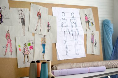 Dikiş odasındaki tahtaya kumaş kalıpları yapıştırılmış. Masanın üzerinde kumaş ruloları ve çok renkli iplikler var. Moda tasarım odası.