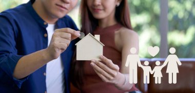 Genç Asyalı adam, tahta ev şeklinde bir para kutusuna gümüş para koyup ikon aileyi gösteriyor. Tasarruf, hayır kurumu, aile finans planı, bağış toplama, yatırım, finansal konsept.