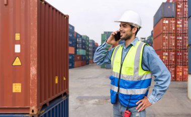 Güvenlik yeleği ve kasketli nakliye işçisi elinde cep telefonuyla ayakta duruyor. Arka planda büyük bir çelik kargo konteynırı var..