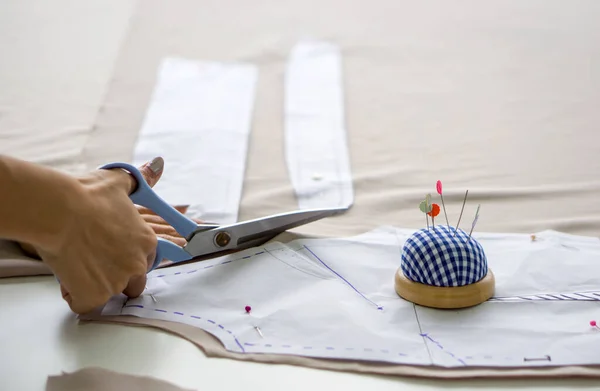Closeup dressmaker hand use scissors to cut fine beige fabric and sketch paper pattern.