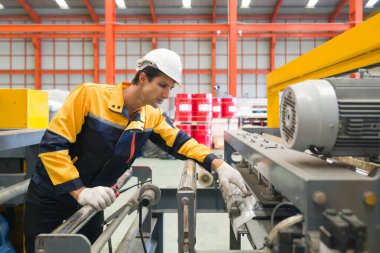 Polyester ceket üniforması, kalın şapka ve koruyucu eldiven giymiş genç İspanyol işçi makinenin düzenini kontrol ediyor. Büyük endüstriyel makinelerle metal levha fabrikasında çalışan bir atmosfer..