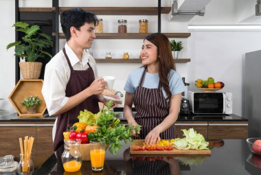 Önlük giymiş genç Asyalı kadın. Domates dilimi, dolmalık biber ve marul. Erkek arkadaşı onun yanında duruyor. Mutfakta sabah atmosferi.