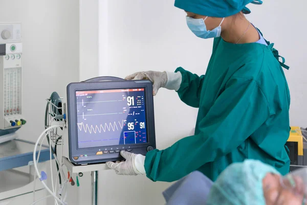 Multi-parameter patient monitor in operating room, controlled by nurse in surgical green gown uniform.