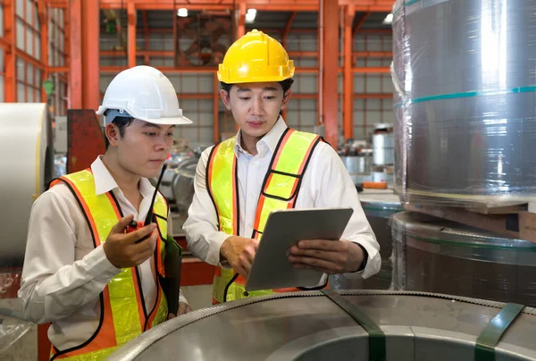 Young asian engineer manager trains new employees within the metal sheet factory. Everyone wear safety vest and hardhat. Steel sheet roll stack are in the working area.