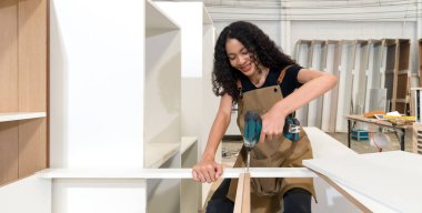 Kahverengi önlüklü genç fabrikada ahşap mobilya monte etmek için pilli kablosuz elektrikli matkap kullanıyor. Çalışma alanına çeşitli boyutlarda ahşap dolaplar konur..