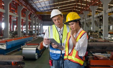 Kıdemli mühendis, metal levha fabrikasında yeni bir çalışan çalıştırıyor. Herkes çelik yelek ve şapka giysin. Çalışma alanında çeşitli endüstriyel makineler var..