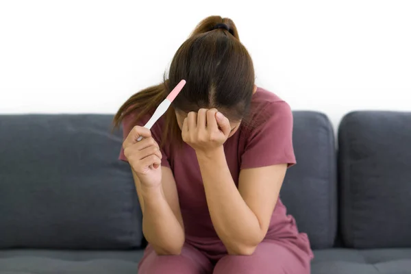 Asian female housewife stressed depressed sitting on sofa in living room holding positive result fertilization pregnancy test kit.