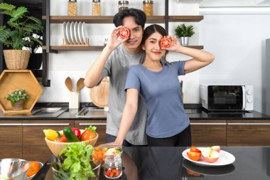 Young couple in casual clothe, hold slice tomato in eye position. Asian lover spend morning time together in the kitchen. 