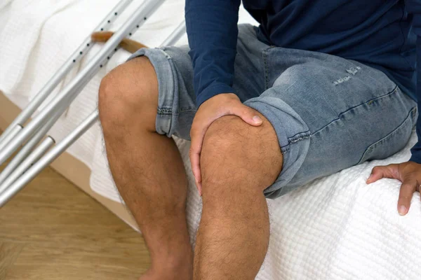 Injured man with painful knee resting on bed in an indoor setting, displaying discomfort. Crutches were placed beside him.