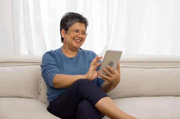 Asian gray hair old woman looking at tablet computer monitor, smiling while sit on sofa in the living room.