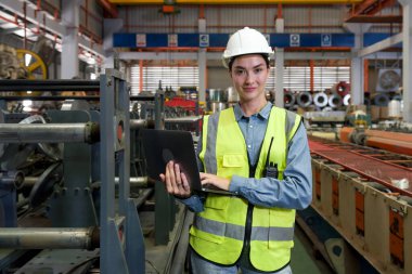 Kendini işine adamış bir kadın mühendis, geniş bir fabrikanın ortasında dizüstü bilgisayar tutuyor..