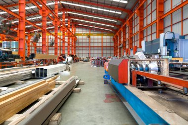 Kocaman bir depo, kafasının üzerinden geçen parıldayan çelik kirişlerden oluşan bir labirentle dolup taşarak metalik bir tepe örtüsü oluşturuyor. Yerde çelik levha ve çelik tüp var..