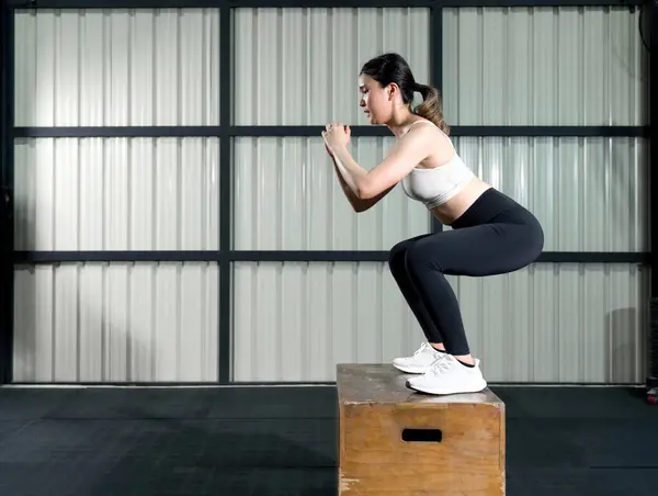 A woman balances atop a sturdy box, her deep concentration visible as she engages in a powerful squat. Her form is strong, sweat glimmers on her face as testament to her perseverance