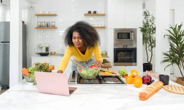 Young woman utilizing digital recipes from laptop computer. Cooking preparation in modern kitchen.