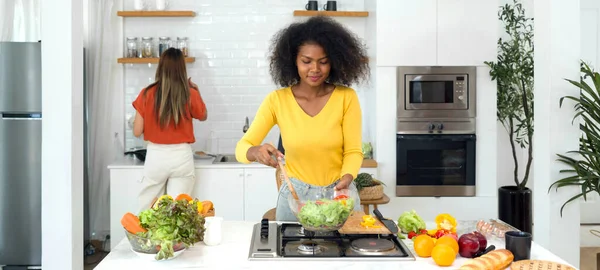 Bustling kitchen scene featuring two women engrossed in food prep. Clad in casual ware, one woman mix salad in glass bowl, the other taste the food in the pan.