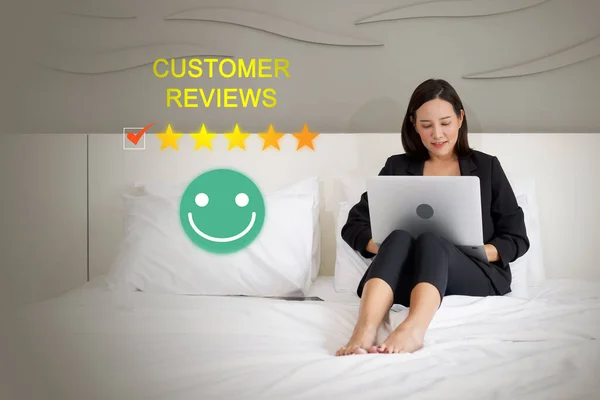 身穿黑色西服的年轻亚洲女商人一边在手提电脑上浏览顾客的评论 一边舒服地坐在酒店房间的床上 — 图库照片