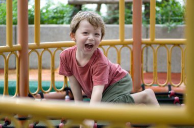 Neşeli genç bir çocuk oyun parkındaki ekipmana tırmanıyor, güneşli gökyüzünün altında açık hava macerasının tadını çıkarıyor..