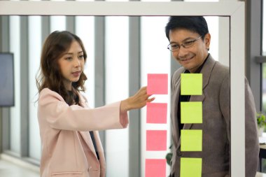 Profesyonel bir erkek ve kadın, şeffaf cam bir duvardaki renkli kağıt notları kullanarak beyin fırtınası yaparak bir araya gelirler..