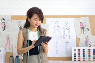 Genç Asyalı terzi tablet bilgisayardan e-posta siparişini kontrol ediyor. Masanın üzerinde kumaş, ipek iplik rengi örnek kataloğu ve çeşitli dikiş malzemeleri var..