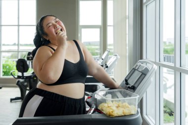 Büyük beden bir kadın spor salonundaki koşu bandında çalışırken cips yiyor, hoşgörü ile fitness 'i birleştiriyor..