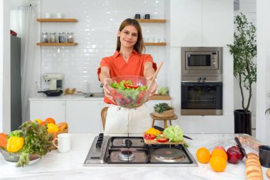 Sağlıklı bir kadın, gülümse ve önüne salata kasesi koy. Modern bir mutfakta sabah atmosferi. Mutfak tezgahı çeşitli sebzelerle dolu..