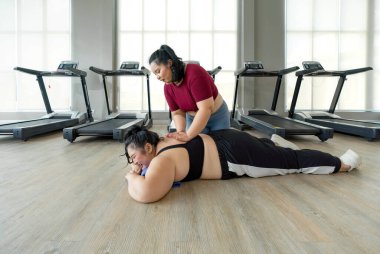 Büyük beden bir bayan koşu bandının yanında dinleniyor, yorgun görünüyor ama iyi. Bir arkadaşı acısını dindirmek için sırtına masaj yapıyor..