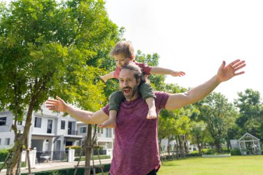 Neşeli bir adam, kıkırdayan küçük bir çocuğu omuzlarında taşır, yemyeşil bir parkta güneşli bir günün tadını çıkarır..